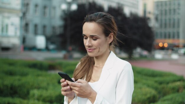 Portret uśmiechniętej profesjonalnej kobiety piszącej wiadomość na telefonie w spacerze po mieście