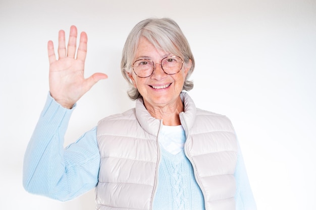 Portret uśmiechniętej siwowłosej starszej kobiety w okularach machającej ręką przed kamerą