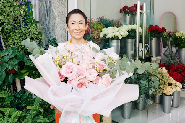 Portret uśmiechniętej starszej właścicielki kwiaciarni stojącej z dużym jasnoróżowym bukietem, który stworzyła dla klienta