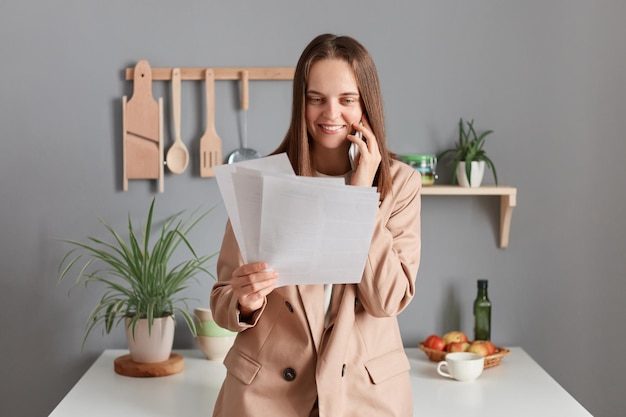 Portret uśmiechniętej szczęśliwej brązowowłosej bizneswoman w beżowym garniturze stojącej przy stole w kuchni w domu, trzymającej dokumenty i rozmawiającej przez smartfon z partnerem biznesowym