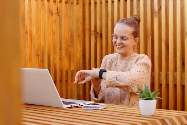 Portret uśmiechniętej, uroczej, zajętej kobiety z fryzurą kok ubraną w beżowy sweter, pracującej na laptopie przy drewnianej ścianie, patrzącej na jej inteligentny zegarek sprawdzający czas, wyrażając optymizm