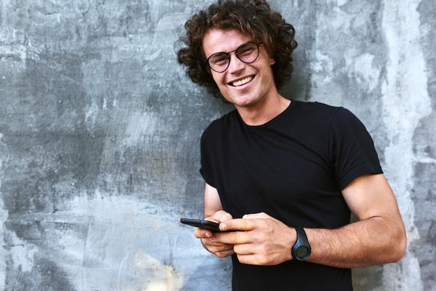 Portret uśmiechnięty mężczyzna stojący na zewnątrz, wysyłając SMS-y na telefon komórkowy, młody mężczyzna z kręconymi włosami nosi okulary, odpoczywając na zewnątrz w mieście, przeglądając jego telefon komórkowy na betonowym szarym tle
