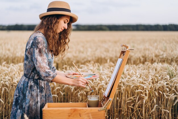 Portret uśmiechnięty żeński artysta z kędzierzawym włosy w kapeluszu. kobieta rysuje obraz krajobrazu na polu pszenicy. Skopiuj miejsce