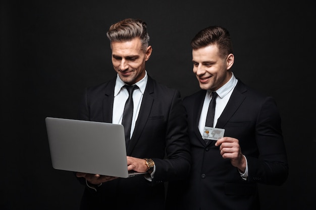 Portret uśmiechniętych dwóch biznesmenów ubranych w formalny garnitur, trzymających laptopa i kartę kredytową na białym tle nad czarną ścianą