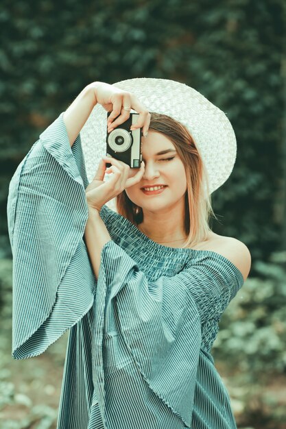 Portret Vintage. Młoda spektakularna kobieta w sukni i kapeluszu robi zdjęcia aparatem retro na niewyraźne tło zewnątrz