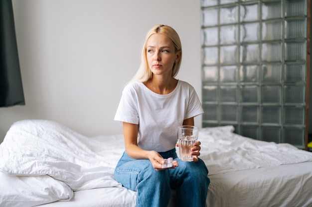 Portret wątpliwej blondynki młodej kobiety trzymającej pigułki i szklankę wody w rękach czuje się niepewnie, biorąc lek