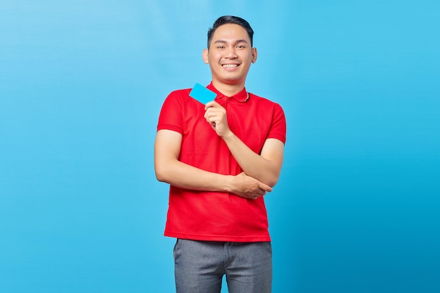 Portret wesołego azjatyckiego młodego mężczyzny pokazującego pustą kartę kredytową i patrzącego na kamerę na białym tle na niebieskim tle