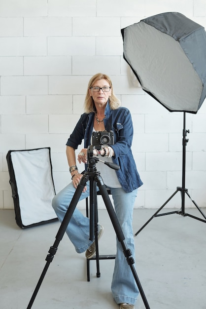 Portret wesołego dojrzałego fotografa w okularach siedzącego podczas sesji zdjęciowej i używającego aparatu