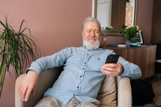 Portret wesołego, dojrzałego mężczyzny trzymającego smartfona za pomocą mobilnej aplikacji online, uśmiechającego się, patrzącego na ekran, siedzącego na wygodnym fotelu Brodaty starszy mężczyzna korzystający z mediów społecznościowych w domu po przejściu na emeryturę