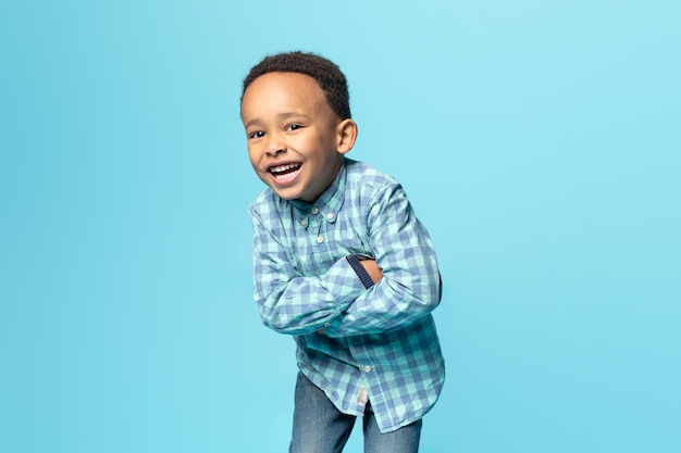 Portret wesołego małego afrykańskiego chłopca pozuje z założonymi rękami i uśmiecha się do kamery na niebieskim tle