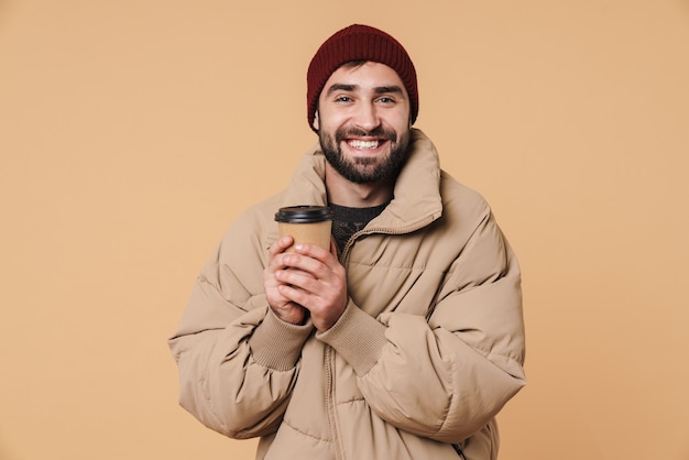 Portret wesołego młodzieńca w zimowej kurtce i kapeluszu uśmiechający się podczas picia kawy na beżowym tle