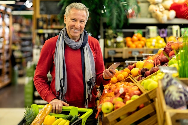 Portret wesołego starszego mężczyzny robiącego zakupy w supermarkecie, trzymającego smartfona