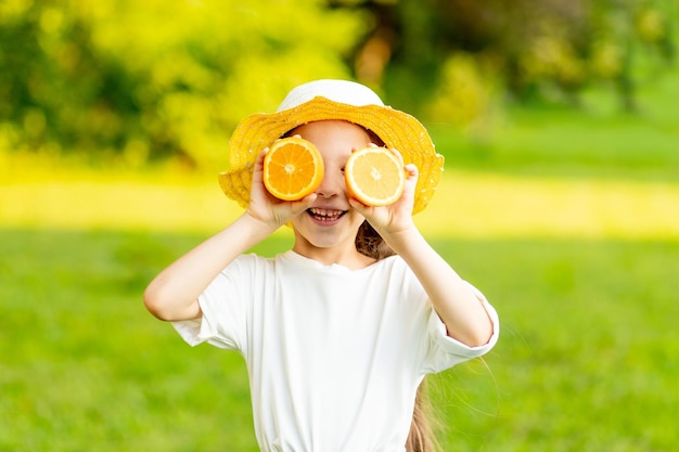 Portret wesołej dziewczynki w żółtym kapeluszu na zielonym trawniku latem uśmiecha się lub śmieje radośnie, zamykając oczy pomarańczą