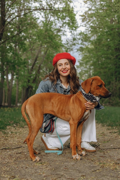 Portret wesołej kobiety pieszczącej brązowego psa Rhodesian Ridgeback w parku i patrzącej na kamerę