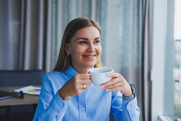 Portret wesołej kobiety w klasycznych okularach uśmiechający się w wolnym czasie w kawiarni z kawą pozytywna europejska kobieta w niebieskiej koszuli na biurku z laptopem zdalną pracę