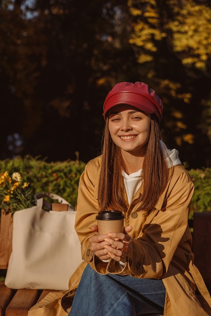 Portret wesołej kobiety w okopie z papierowym kubkiem kawy na wynos siedzącej na ławce w parku