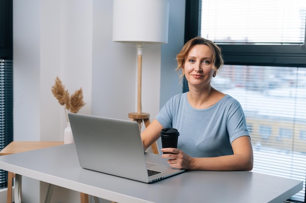 Portret wesołej, ładnej blondynki pracującej na laptopie, trzymającej filiżankę z kawą, patrzącej na kamerę siedzącą przy stole na tle dużego okna