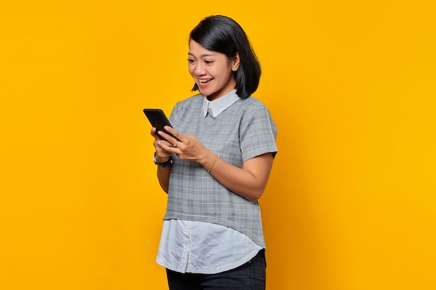 Portret wesołej młodej azjatyckiej kobiety patrzącej na wiadomość na smartfonie na białym tle na żółtym tle