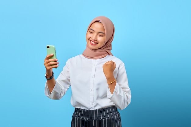 Portret wesołej młodej azjatyckiej kobiety trzyma telefon komórkowy i świętuje sukces z podniesioną ręką