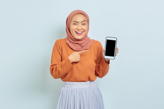 Portret wesołej młodej azjatyckiej muzułmańskiej kobiety w brązowym swetrze i hidżabie, wskazując na pusty ekran telefonu komórkowego na białym tle muzułmański styl życia koncepcja