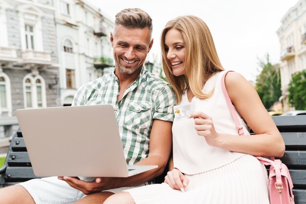 Portret wesołej młodej pary w letnie ubrania, trzymając kartę kredytową i korzystając z laptopa, siedząc na ławce na ulicy miasta