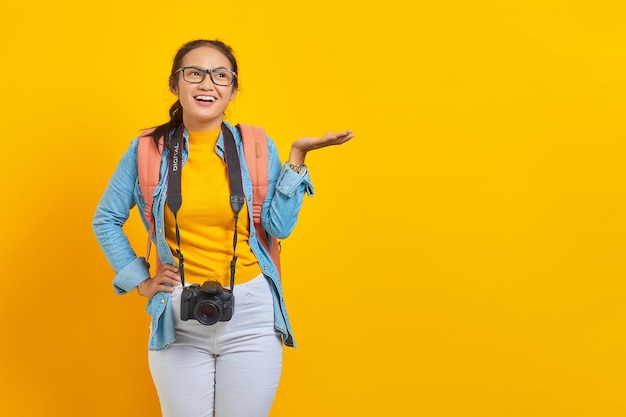 Portret wesołej młodej podróżniczki azjatyckiej kobiety z plecakiem i aparatem w dżinsowych ubraniach, zapraszając na żółtym tle. Pasażer podróżujący w weekendy. Koncepcja podróży samolotem