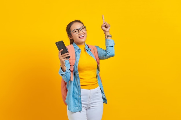 Portret wesołej młodej studentki Azjatki w swobodnych ubraniach z plecakiem trzymającym telefon komórkowy i wskazującą w górę przestrzeń palcami na białym tle na żółtym tle