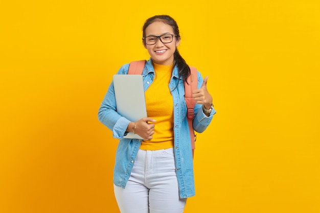 Portret wesołej młodej studentki azjatyckiej kobiety w zwykłych ubraniach z plecakiem trzymającym laptopa i pokazującym kciuki do góry z palcem na białym tle na żółtym tle Edukacja w koncepcji college'u