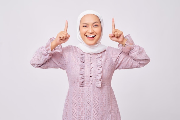 Portret wesołej, pięknej, młodej azjatyckiej muzułmanki noszącej hidżab i fioletową sukienkę wskazującą palcem w wolne miejsce na reklamę odizolowaną na tle białego studia