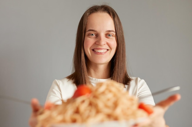 Portret wesołej, zachwyconej kaukaskiej kobiety o ciemnych włosach, która bierze pyszny makaron, jedząc obiad odizolowany na szarym tle, będąc głodną, uśmiechając się radośnie