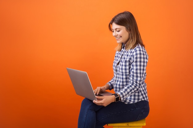 Portret wesołej zajętej kobiety z brązowymi włosami w kraciastej casualowej koszuli i dżinsowej siedzącej z zębatym uśmiechem, pracującej na laptopie, pisząc na klawiaturze, studio strzał na białym tle na pomarańczowym tle