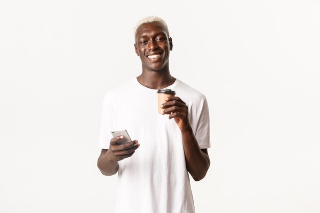 Portret wesoły afroamerykański mężczyzna pije kawę, używa telefonu komórkowego i uśmiecha się szczęśliwy do kamery,