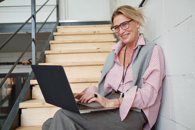 Portret wesoły dojrzały bizneswoman siedzi na schodach i za pomocą laptopa podczas przygotowywania raportu sprzedaży