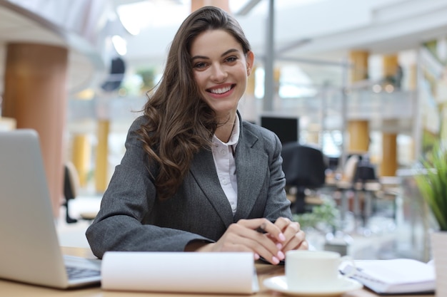 Portret wesoły młody bizneswoman siedzi przy stole w biurze i patrząc na kamery.