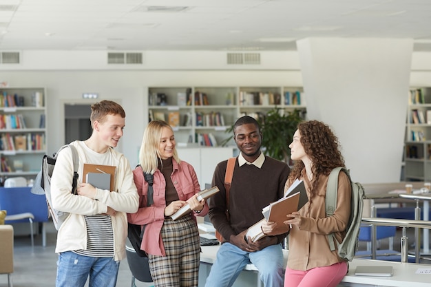 Zdjęcie portret wieloetnicznej grupy studentów stojących w bibliotece uczelni i rozmawiających, trzymając książki i plecaki, powyżej