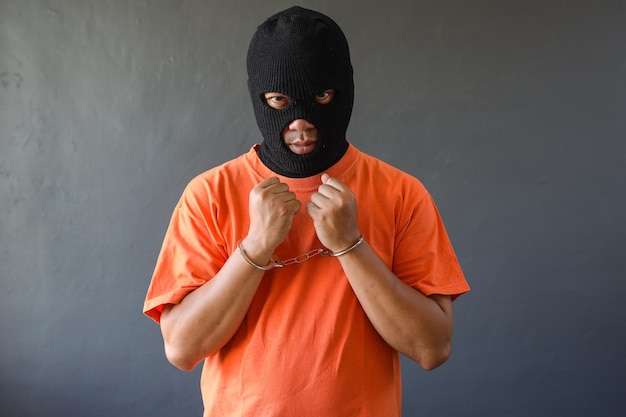 Portret więźnia w czarnej masce i pomarańczowej koszulce w kajdankach odizolowanych na szarym tle