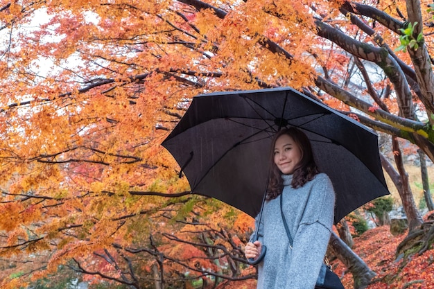 Portret wizerunek pięknej azjatyckiej kobiety stojącej w deszczu z czerwonymi i pomarańczowymi liśćmi drzewa jesienią w tle