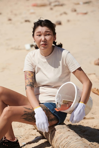 Portret wolontariuszki odpoczywającej na pniu drzewa na plaży po zebraniu śmieci w plastikowym wiadrze