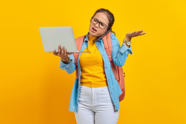 Portret wściekłej młodej studentki azjatyckiej kobiety w zwykłych ubraniach z plecakiem rozmawiającej na smartfonie, trzymając laptopa, mówiąc „nie” z dłońmi na białym tle na żółtym tle