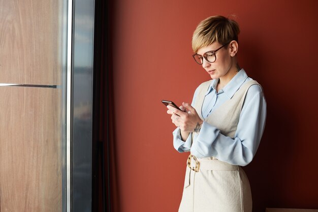 Portret współczesnej krótkiej bizneswoman za pomocą smartfona, stojąc przed czerwoną ścianą w biurze, miejsce