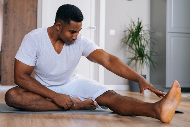 Portret wysportowanego Afroamerykanina ćwiczącego jogę, siedzącego w ćwiczeniu Janu Sirsasana, w pozycji Head to Knee Forward Bend, na podłodze podczas ćwiczeń w jasnym salonie.