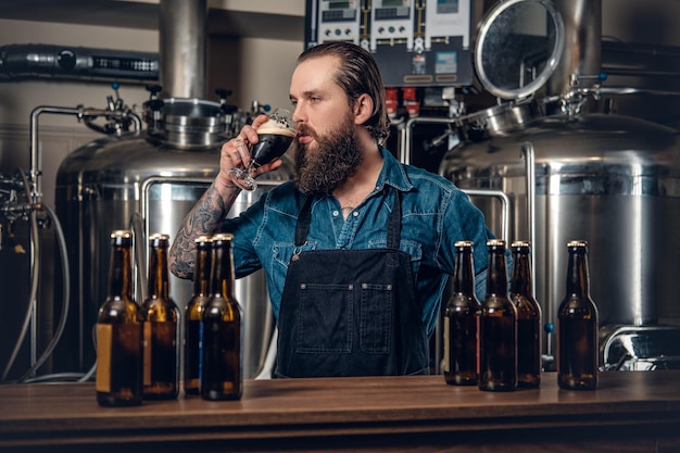 Portret wytatuowanego, brodatego męskiego producenta hipstera degustującego piwo w minibrowarze.