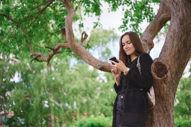 Portret z dolnego kąta młodej kobiety w czarnej kurtce ze smartfonem w dłoniach na tle dużego rozgałęzionego drzewa. komunikacja wirtualna, generacja Z
