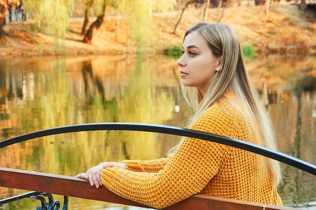 Portret z profilu zamyślonej blondynki w żółtym swetrze relaksuje się w jesiennym parku w pobliżu jeziora