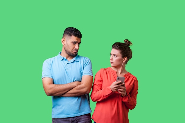 Portret zabawnego, urażonego mężczyzny w swobodnym stroju stojącego ze skrzyżowanymi rękami, patrzącego z dezaprobatą na kobietę trzymającą telefon komórkowy uzależnioną od Internetu na zielonym tle strzał w studio