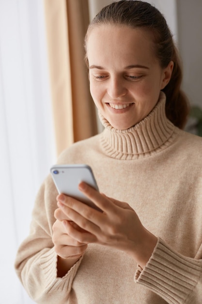 Portret zachwycona młoda dorosła kobieta z fryzurą w kucyk, ubrana w beżowy sweter w stylu casual, korzystająca z telefonu komórkowego, pisząca posty na portalach społecznościowych lub oglądając zdjęcia.