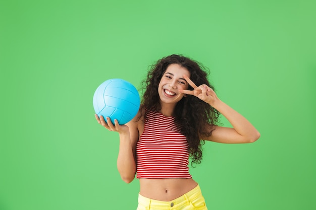 Portret zachwyconej kobiety w wieku 20 lat w letnich ubraniach, uśmiechniętej i trzymającej piłkę do siatkówki, stojąc na zielono