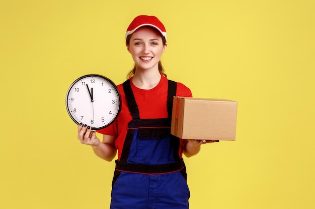 Portret zachwyconej kurierki kobiety stojącej z kartonową paczką i zegarem ściennym dostarczającym paczkę do klienta na czas w kombinezonie i czerwonej czapce Kryty strzał studio na białym tle na żółtym tle