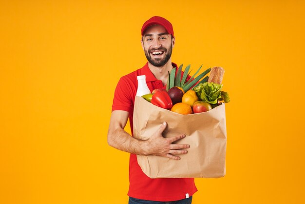 Portret zadowolonego dostawcy w czerwonym mundurze uśmiechającego się, niosąc papierową torbę z produktami spożywczymi izolowanymi na żółto