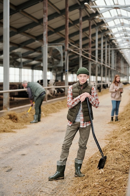 Portret zadowolonego nastoletniego chłopca w kapeluszu stojącego z łopatą w oborze, który pracuje z rodzicami na farmie